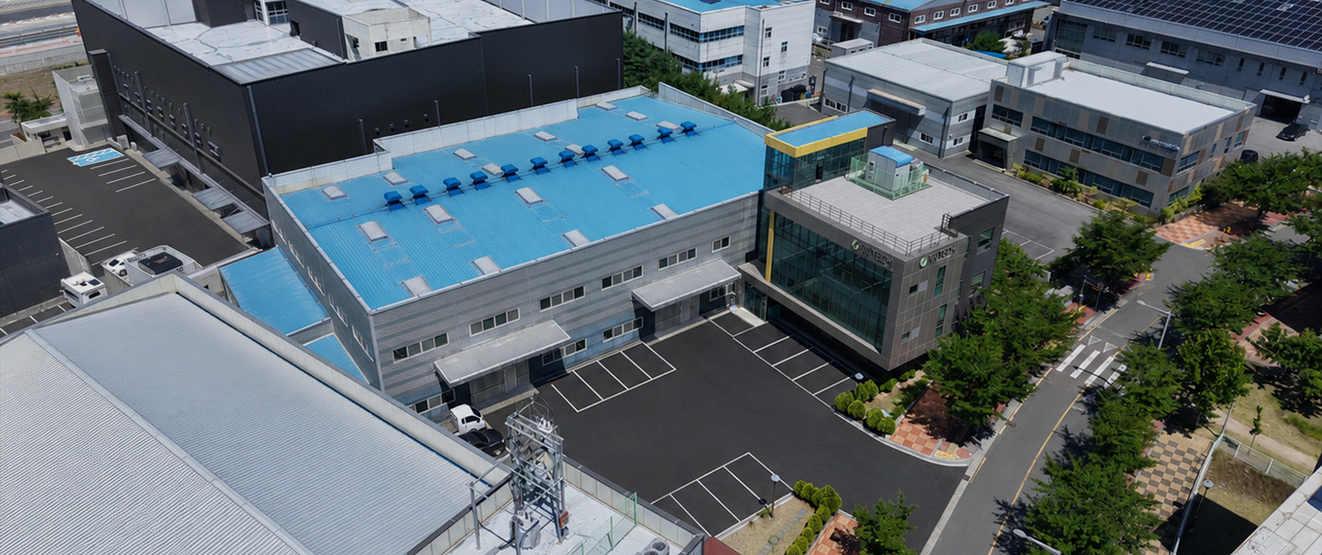 Aerial top-down view of the company campus including blue-roofed manufacturing facilities, office building, and parking lot